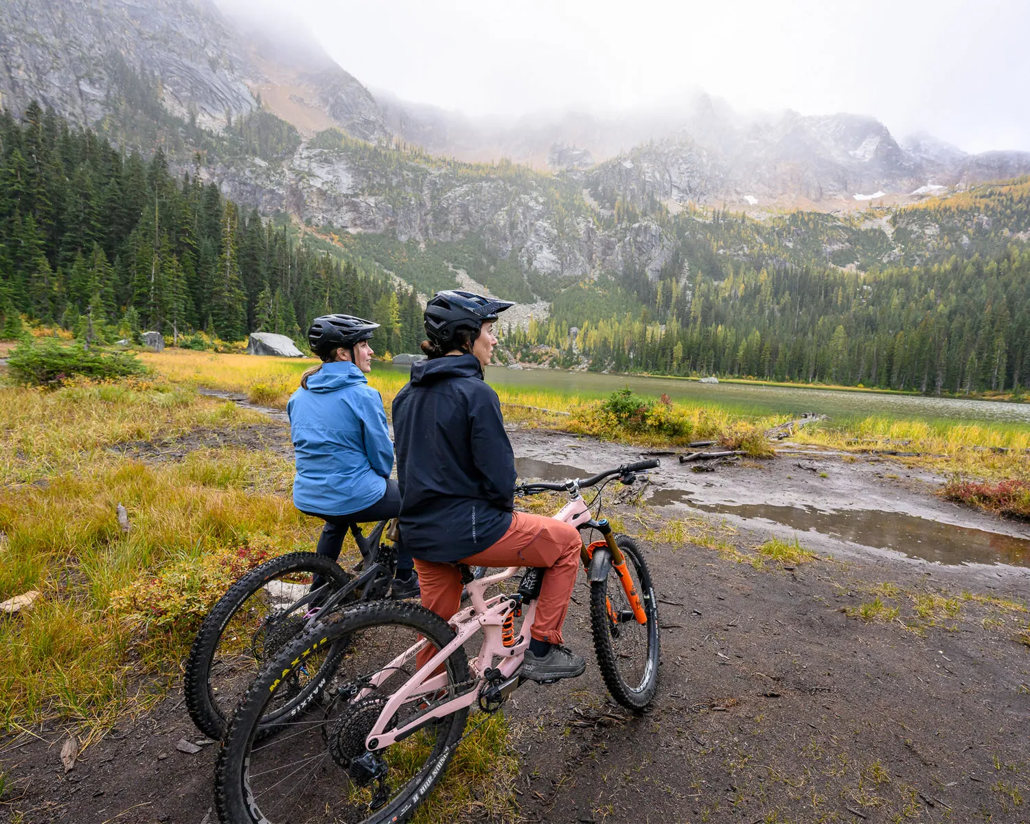 Two mountain bikers take in a view of the mountains while sitting on their bikes on a wet, muddy day.